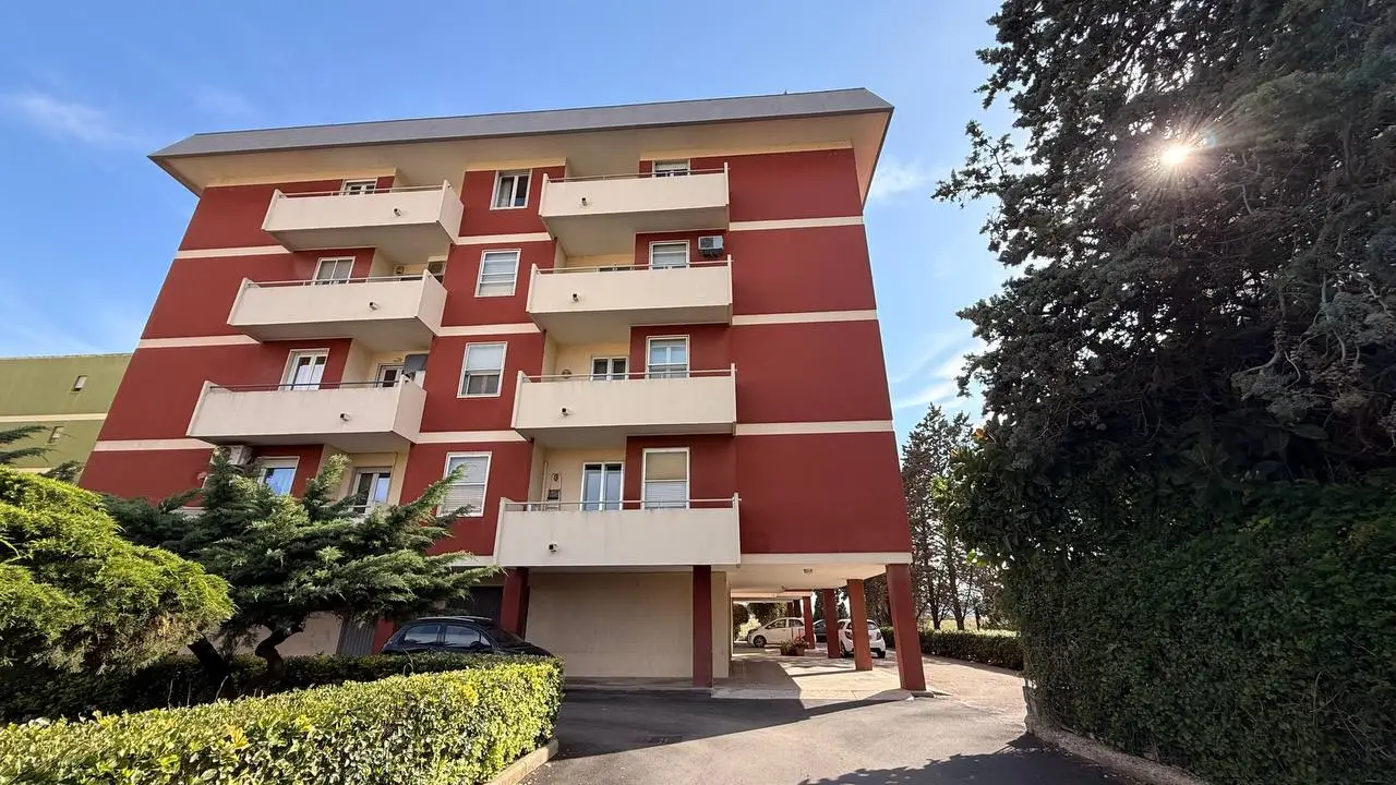 Palazzina residenziale a più piani con facciata rossa e balconi bianchi, circondata da alberi e area parcheggio privata.